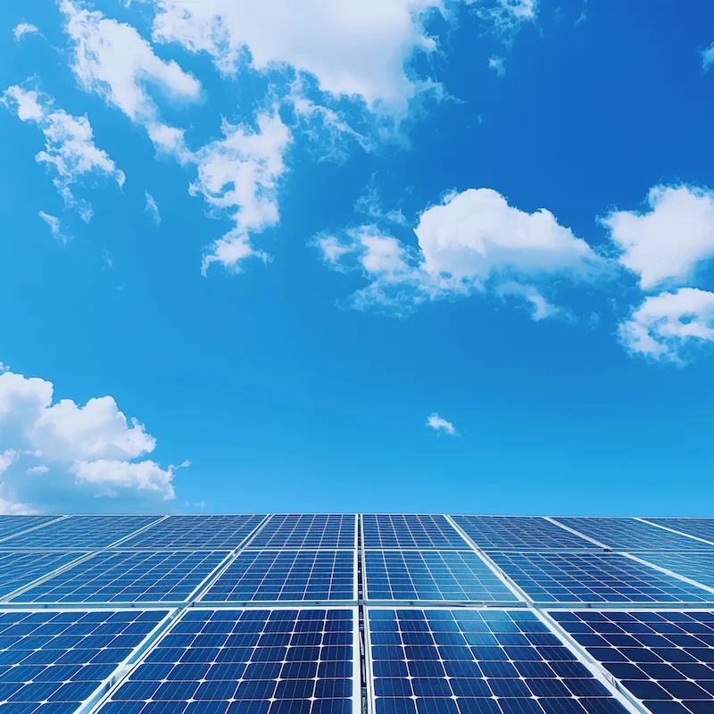 A vibrant image displaying a large array of solar panels under a clear blue sky with scattered white clouds. The solar panels are positioned to capture maximum sunlight, illustrating sustainable energy production. This scene symbolises the integration of renewable energy technologies with natural environments to harness clean power and reduce air pollution.